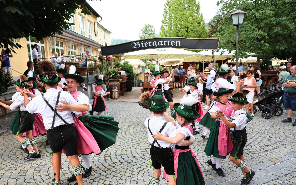 Mädchen und Jungen in traditionellen bayerischen Trachten tanzen auf dem Marktplatz in Berchtesgaden vor dem Biergarten des Gasthof NEUHAUS, während zahlreiche Zuschauer zuschauen.