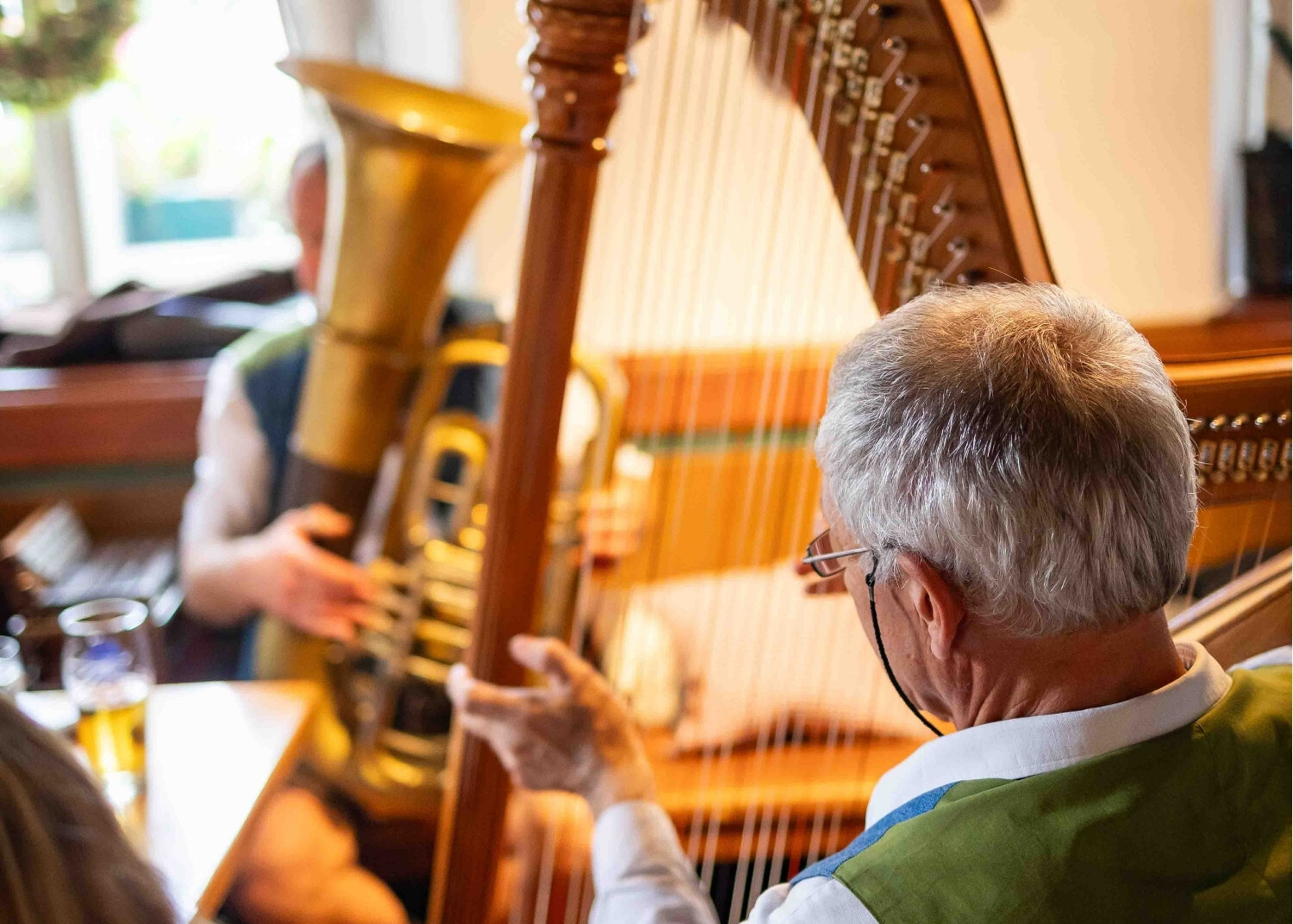 An older man with gray hair and glasses plays a large wooden harp, while another person in the background holds a trumpet, in a cozy room filled with natural light.
