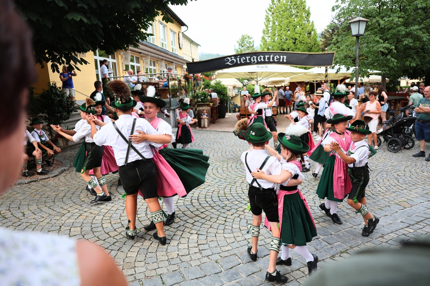Children dancing in traditional Bavarian costumes in front of a beer garden, surrounded by spectators and visitors.