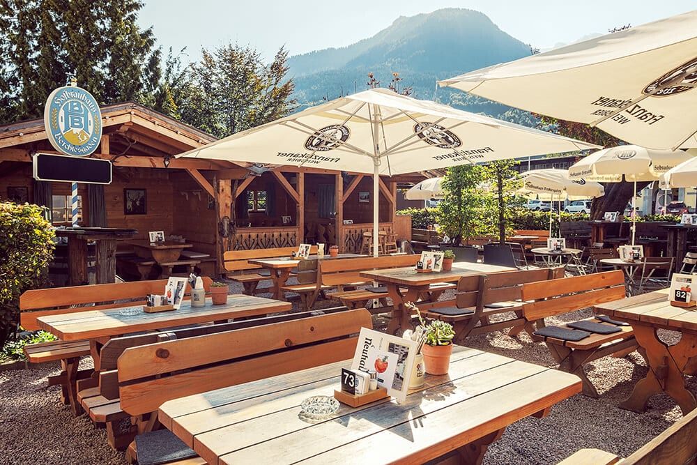 Ein outdoor Restaurantbereich mit mehreren Holztischen und Bänken, großen Sonnenschirmen, einer kleinen Holzhütte im Hintergrund, und bergigem Landschaftshintergrund.