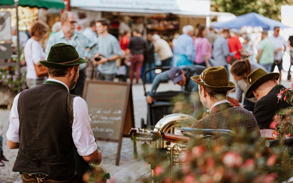 Drei Männer mit traditionellen grünen Filzhüten, einer trägt eine schwarze Weste und weiße Hemd, sitzen beim Musizieren auf einem Volksfest, umgeben von einem bunten Menschenmengen im Hintergrund.
