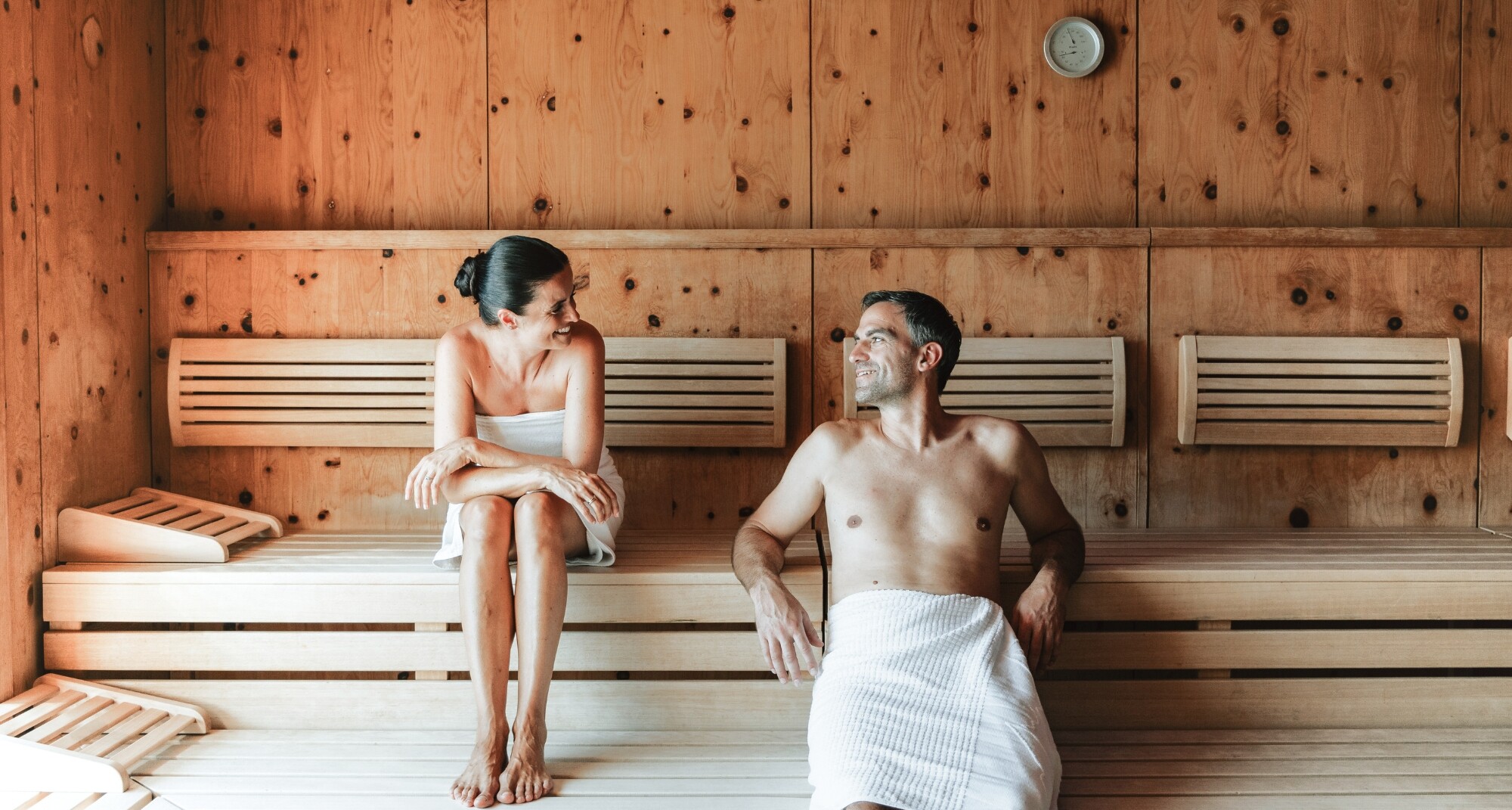 Ein Paar sitzt in einer Holz-Sauna, eine steht auf der Bank und lächelt die andere an, die auf dem Boden sitzt und ihre Beine umarmt, beide tragen Handtücher.