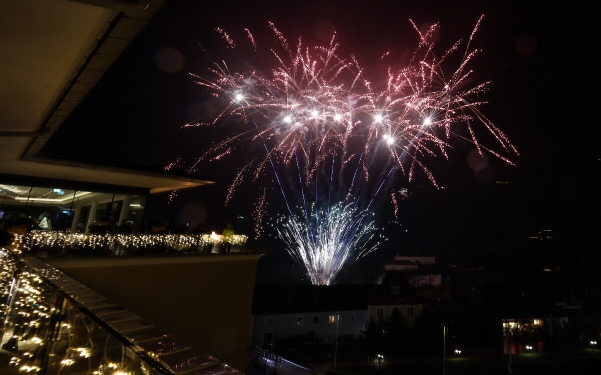 Feuerwerksraketen explodieren in der Nacht, mit leuchtenden Funken und Lichtern am Himmel, sichtbar von einem Balkon oder einer Terrasse mit beleuchteter Geländer in einer Stadt.