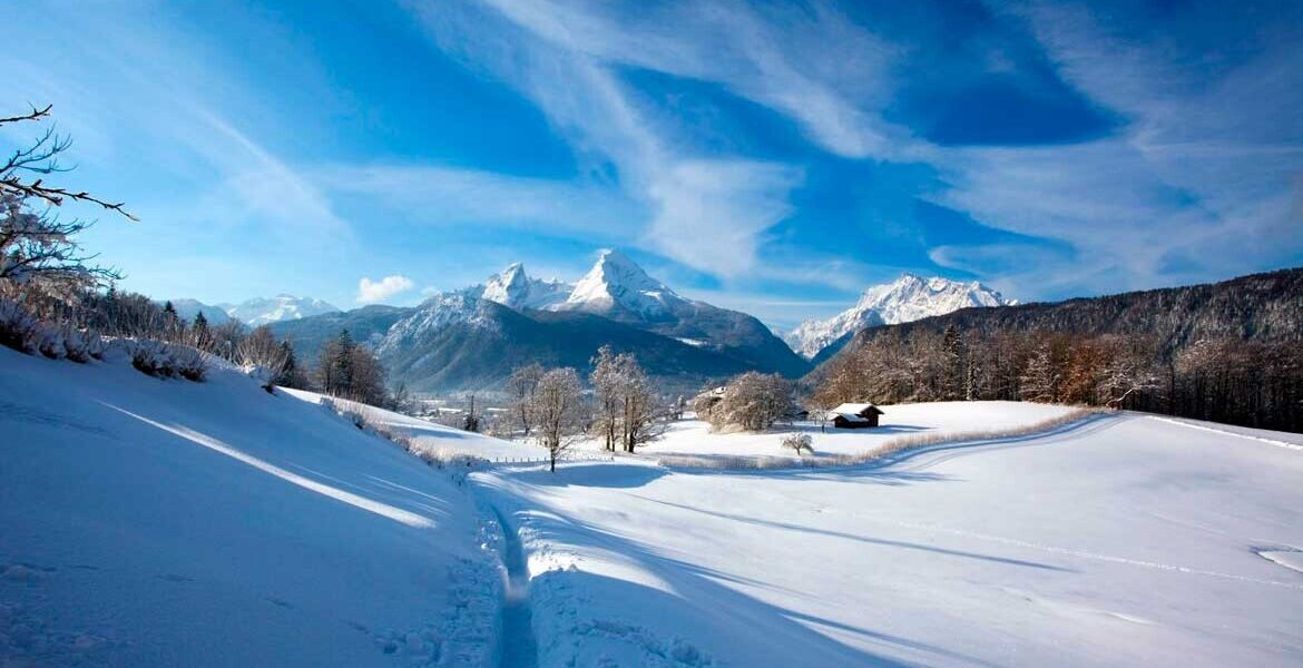 Berglandschaft mit verschneiten Feldern, Bäumen und Bergen im Hintergrund, unter einem blauen Himmel mit Wolken.