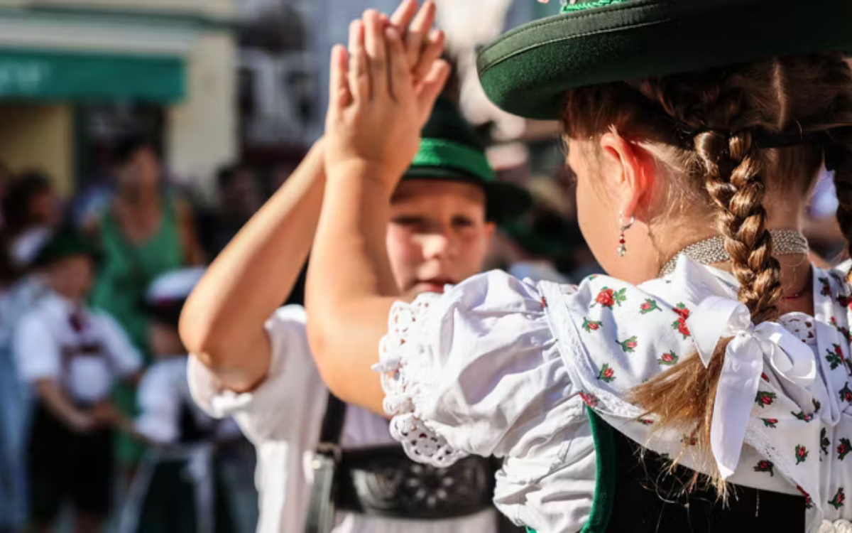 Zwei Kinder in traditioneller Kleidung beim Händeschütteln bei einem Fest, im Hintergrund weitere Kinder und Erwachsene.