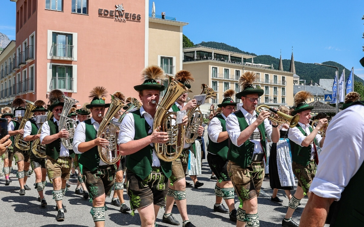 Männer in traditioneller Tracht mit Hut und Feder schmuck spielen Alfonsorchester während eines Festumzugs auf der Straße, im Hintergrund das Hotel EDELWEISS Berchtesgaden und ein bergiges Gelände.