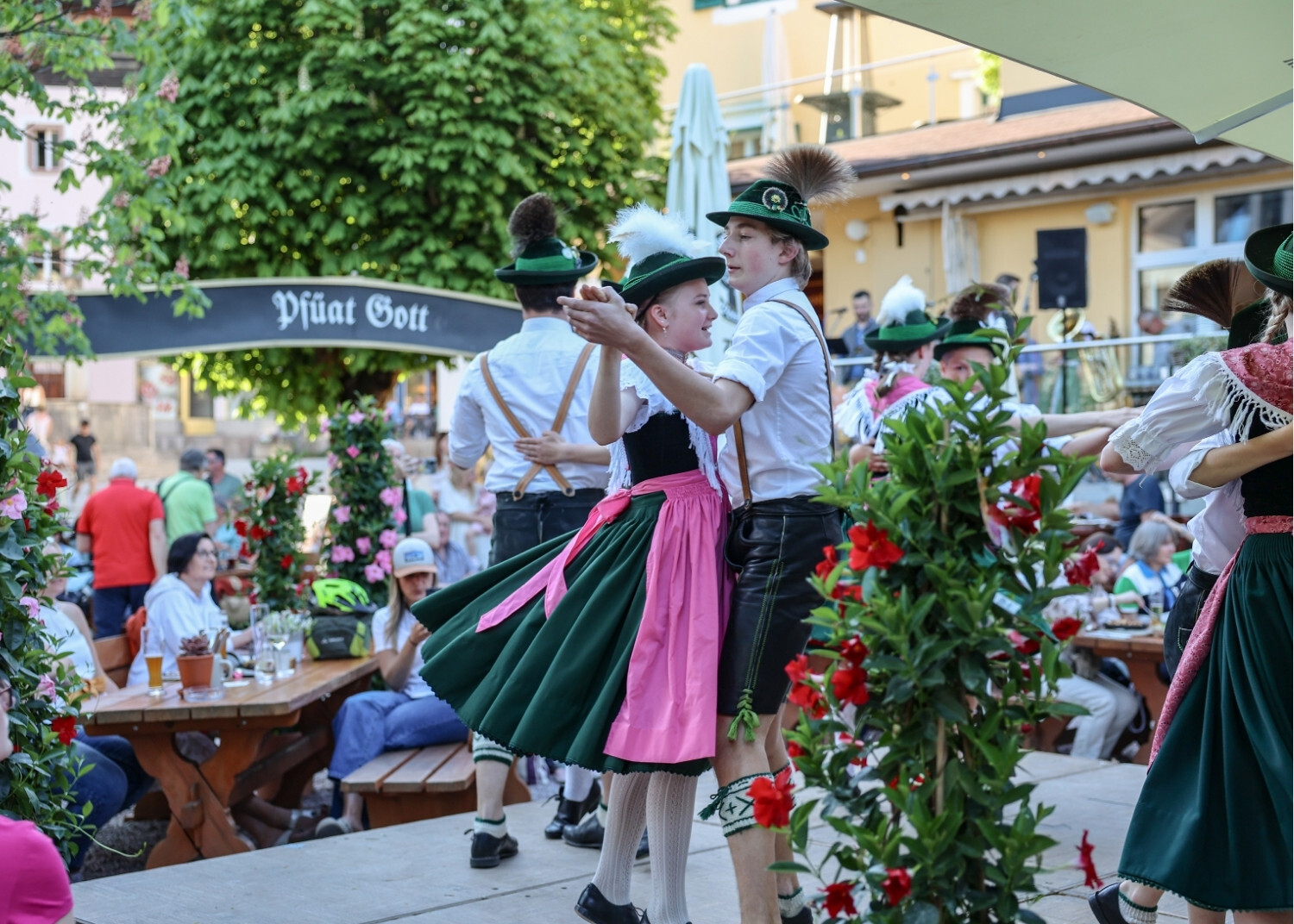 Paare in traditioneller bayerischer Kleidung tanzen auf einer Bühne bei einem Fest, umgeben von Blumen und einheimischen Zuschauern.