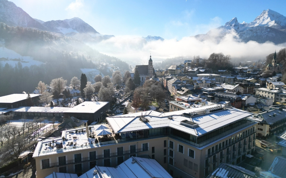 Luftaufnahme eines verschneiten Dorfes in den Bergen mit alten und modernen Gebäuden, umgeben von schneebedeckten Bäumen und hohen, schroffen Alpen im Hintergrund, bei bewölktem Himmel mit Sonnenlicht.