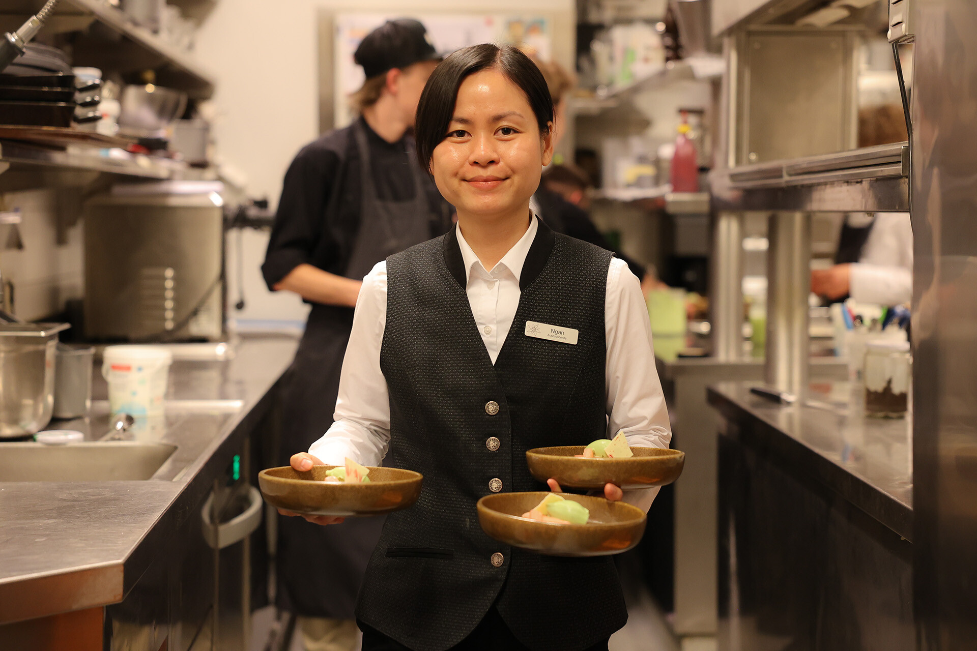 Service-Auszubildende trägt Vorspeisen aus der Küche, im Hintergrund arbeiten die angehenden Köche – praxisnahe Hotellerie-Ausbildung und Teamwork am Lehrlingstag 2025