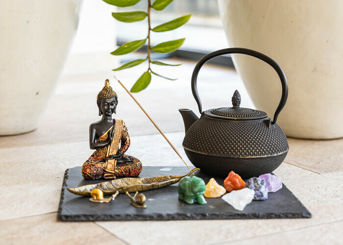 Eine schwarze Teekanne, eine sitzende Buddha-Figur, mehrere bunten Kristalle, eine kleine grüne Statue eines Tieres, und eine goldene Schnecke auf einem schwarzen Steinuntergrund, umgeben von Pflanzen im Hintergrund.