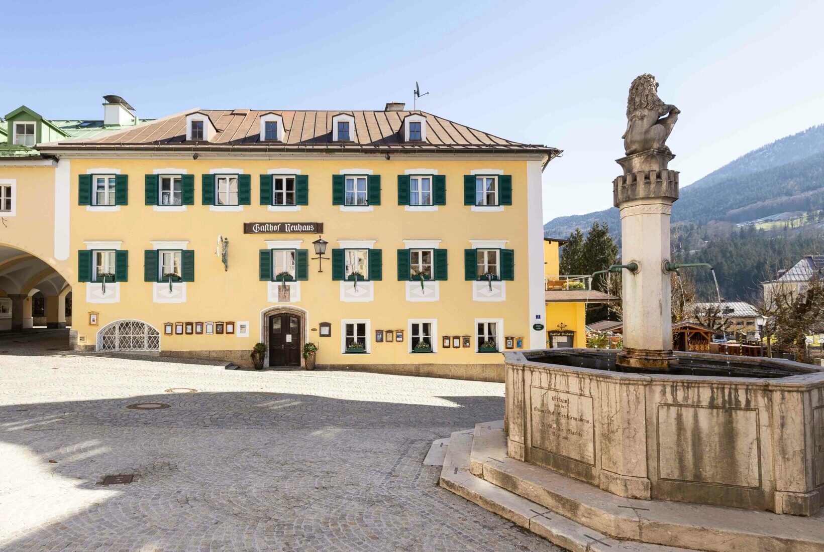 Ein gelb gestrichenes Gebäude mit grünen Fensterläden und schwarzem Eingang, daneben eine alte Brunnen mit Skulptur, in einer gepflasterten, offenen plaz, im Hintergrund Berge.