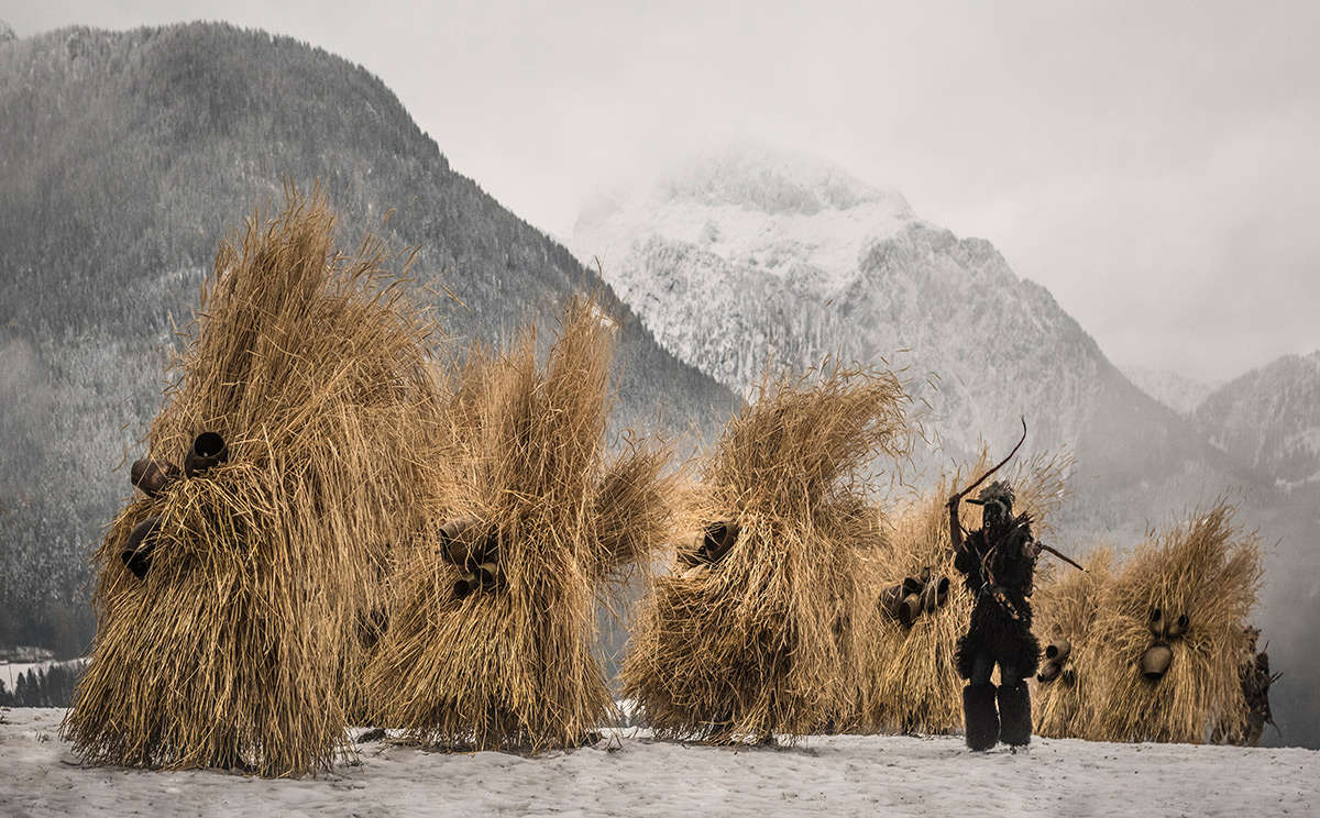 Menschen in traditionellen Kostümen tragen große Heuhaufen auf dem Rücken, während sie auf verschneitem Boden vor einer bergigen, winterlichen Landschaft stehen.